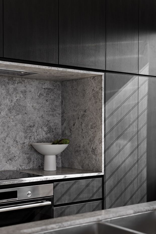 Close-up of a sophisticated dark-toned kitchen featuring vertically grained charcoal cabinetry, a grey natural-stone benchtop and open stone-lined niche, integrated stainless-steel oven, and a partially visible sink in the foreground. Strong diagonal sunlight creates graphic shadows across the cabinetry.