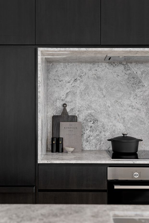 Close-up of a sophisticated dark kitchen with near-black timber cabinetry, a pale grey heavily veined stone splashback and matching recessed benchtop niche. A black cooktop pot, dark chopping board, small bowl and countertop oven are visible, with a stainless-steel oven integrated below. The composition is moody, monochromatic and refined.