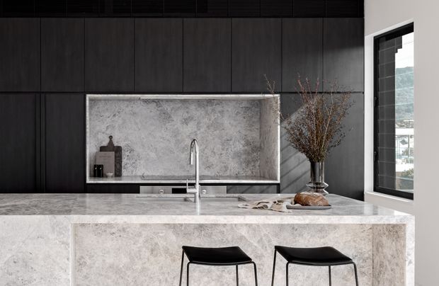 A contemporary kitchen with a large pale stone island and matching raised splashback, dark flat-panel cabinetry, a minimalist stainless-steel tap, and two black bar stools. A window provides natural light and views beyond.
