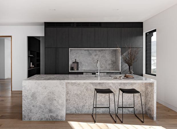 A refined contemporary kitchen featuring a large stone island with waterfall ends and two black bar stools, a matching stone splashback, and extensive dark vertical-panel cabinetry. A sink and minimalist tap sit beneath the splashback, with open shelving to the side and timber flooring throughout. Natural light enters through a large window on the right.