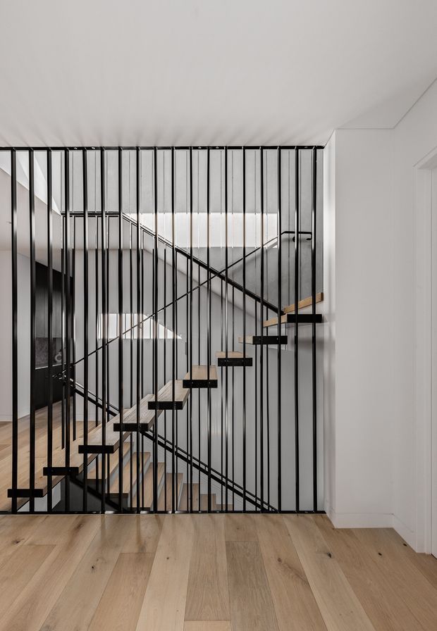 A contemporary interior stairway viewed through a striking black metal vertical-bar screen. The open-riser staircase has light timber treads and black steel framing, with a second flight and landing visible behind it. Pale timber flooring, white walls and ceiling, and strong linear geometry create a clean, minimalist composition.