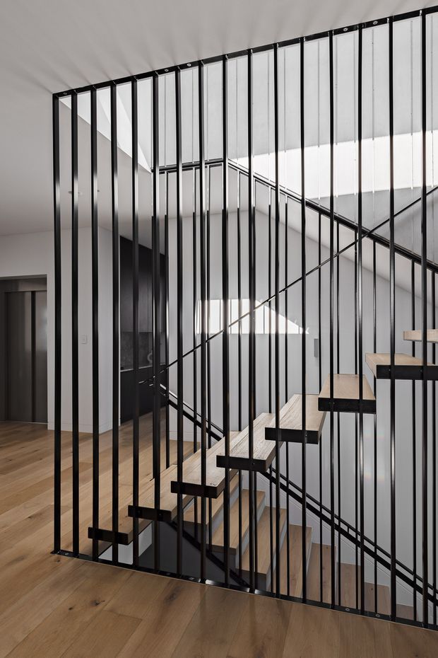 A contemporary interior stairway viewed through a striking black metal vertical-bar balustrade. The open floating staircase has pale timber treads, dark structural supports, and rises through a bright multi-level stair void. Warm timber flooring contrasts with white walls and ceilings, while daylight enters through high-level glazing or a clerestory.