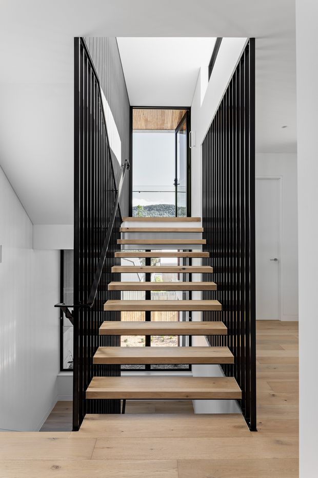 A contemporary interior view of an open-riser staircase with floating light timber treads, slim black metal framing, and vertical black slat balustrades. The stairwell connects multiple levels and is illuminated by a tall glazed opening, with pale timber flooring, white walls, and a glimpse of the surrounding landscape beyond.