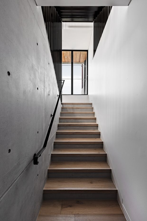 A narrow modern stairway rises between a raw concrete wall and a smooth white wall. The staircase has warm timber treads with dark risers, a slim black handrail, and black vertical slatted screening overhead and along the upper landing. Glazed doors at the top open toward a bright exterior or balcony, with a timber-lined ceiling visible beyond.