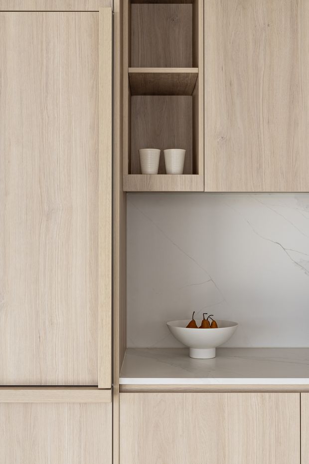 Close-up of a refined built-in timber kitchen joinery composition. Pale oak-toned cabinetry surrounds an open display niche with darker timber backing and shelving, while a light veined stone backsplash and matching shelf surface frame a ceramic bowl of pears. The design is warm, minimal, and highly tailored, with clean flush fronts and understated detailing.