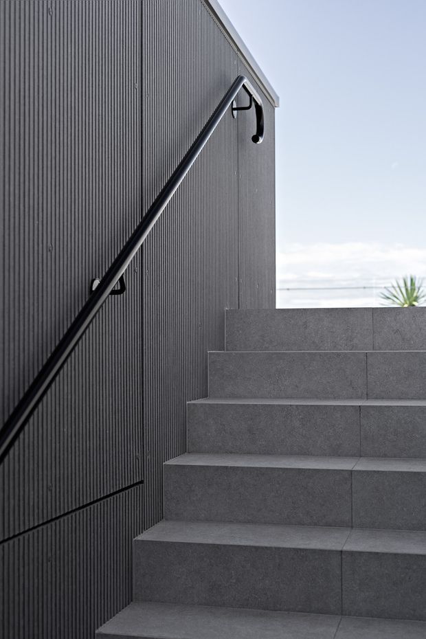Close-up exterior view of a contemporary stairway with broad grey tiled steps, a slim black handrail, and a dark vertically ribbed wall. The stair rises beside a minimalist building facade, with bright sky and a small amount of coastal-looking background visible beyond.