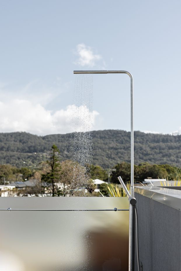 An outdoor shower with a slender brushed-metal frame and overhead rainfall outlet, positioned beside a frosted privacy screen and a dark tiled or rendered wall. Water is visibly running, with a leafy hillside and residential landscape in the background under a pale blue sky.
