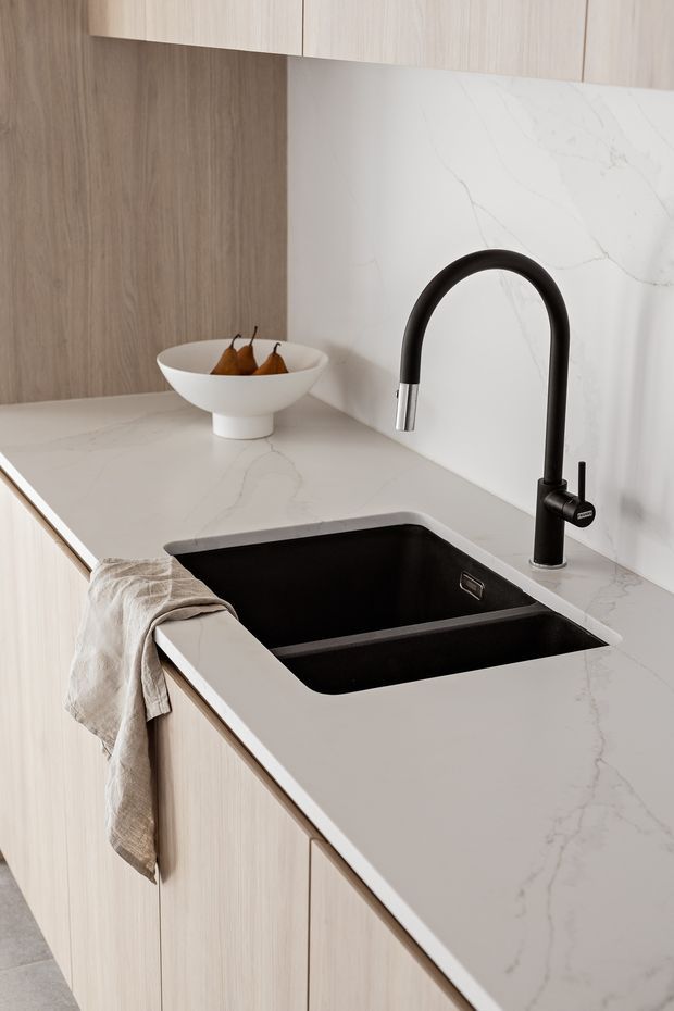 Close-up of a refined contemporary kitchen featuring pale timber cabinetry, a white veined stone benchtop and splashback, and a large black undermount double-bowl sink with a matte black and chrome gooseneck tap. A ceramic fruit bowl and linen tea towel add subtle styling.
