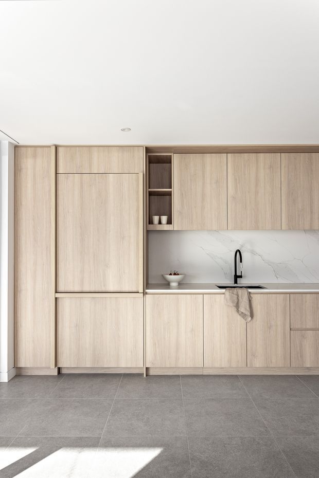 A minimalist contemporary kitchen with full-height light timber-grain cabinetry, integrated storage, a white marble-look splashback with subtle veining, a slim dark benchtop, and a black gooseneck tap. Large grey floor tiles and a white ceiling create a clean, restrained interior.