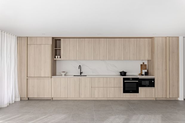 A clean, contemporary galley-style kitchen with a full wall of light timber cabinetry, integrated black appliances, a pale marble-look splashback, and a grey tiled floor. Sheer curtains line the left side, while minimal countertop styling and concealed storage create a calm, streamlined appearance.