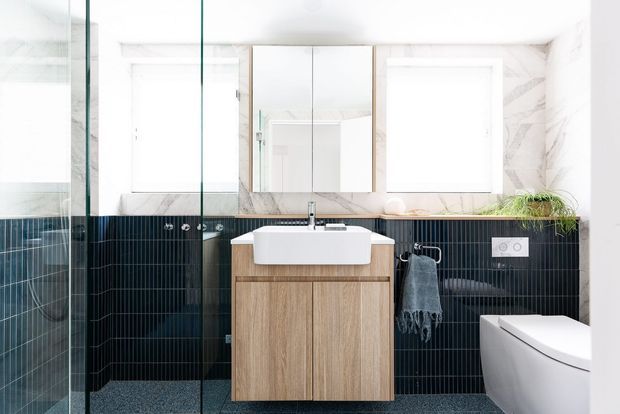 Bright contemporary bathroom with a floating light-wood vanity, white vessel basin, mirrored cabinet, frameless glass shower enclosure, white toilet, and dark vertically ribbed wall tiles. White marble-look surfaces and large windows create a clean, high-contrast interior.