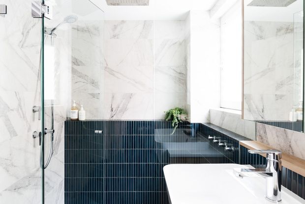 Bright contemporary bathroom featuring large white marble-look wall slabs, dark vertically ribbed wall tiles, a frameless glass shower, wall-mounted basin with chrome tapware, timber vanity shelf, and a large mirror. Natural light enters through the window, creating a clean, refined atmosphere.