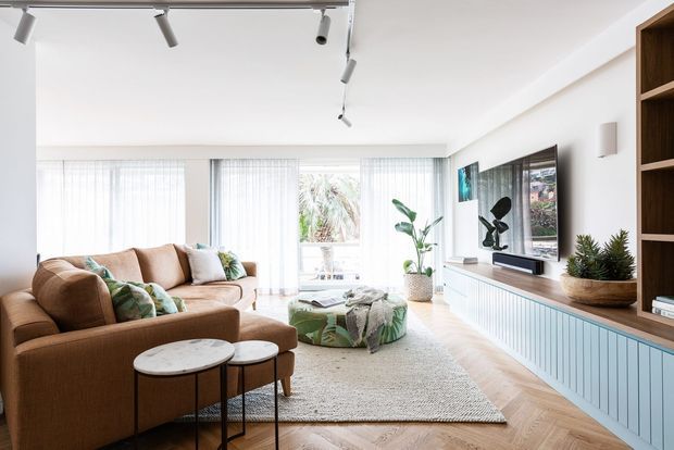 Bright contemporary living room with a large tan sectional sofa, patterned ottoman, nested round coffee tables, pale textured rug, indoor plants, and a wall-mounted television. The room features white walls, full-height sheer window coverings, herringbone timber flooring, a built-in ribbed light-blue media cabinet, and open timber shelving. Track lighting runs across the ceiling, and a tropical garden is visible through the glazing.
