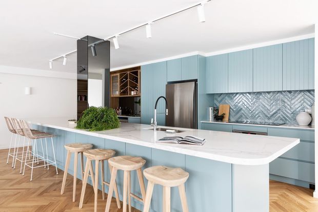 Bright contemporary kitchen with extensive pale blue vertical-panel cabinetry, a large white waterfall island and integrated breakfast bar, stainless steel refrigerator, and a grey-blue herringbone tile splashback. Timber bar stools and a warm parquet timber floor contrast with the cool cabinetry. A built-in timber wine display and open shelving sit beside a dark central structural or service element, with track lighting across the white ceiling.
