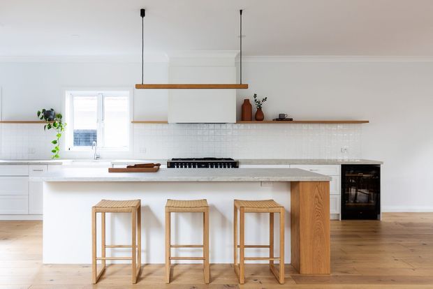 Bright, minimalist white kitchen with a long island, integrated cooktop, white tiled splashback, timber cabinetry detail, and three woven timber bar stools. Light timber flooring, open shelving, pendant lighting, and sparse decorative styling create a warm contemporary Scandinavian feel.