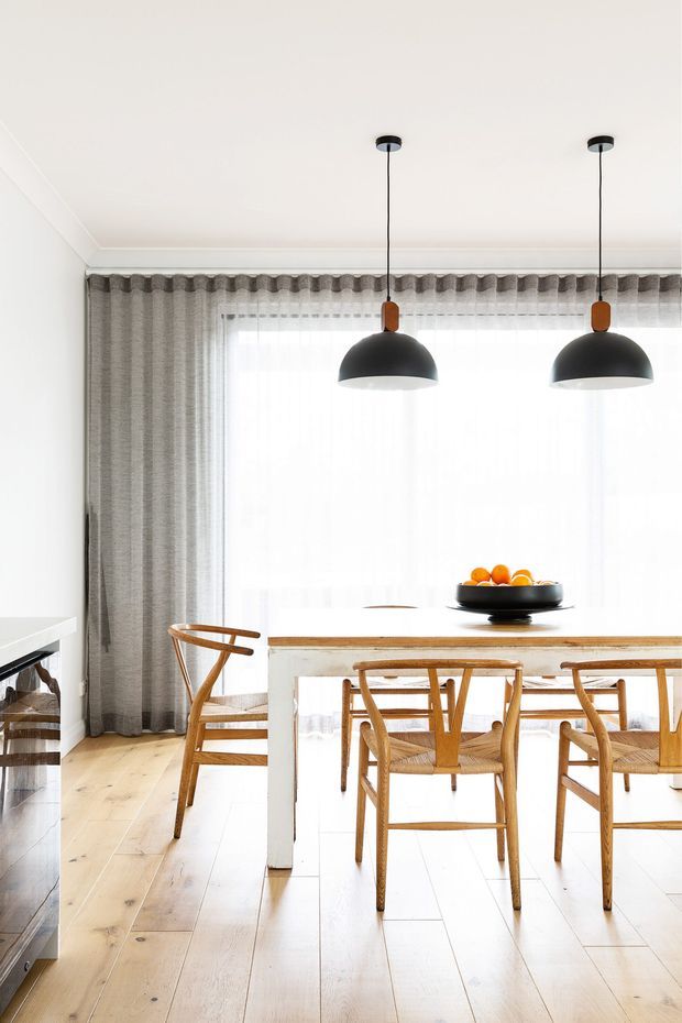 Bright, refined dining area with a white-painted ceiling and walls, pale timber flooring, a simple white dining table with timber top, and woven timber dining chairs. Full-height grey curtains frame a large bright window, while two black dome pendant lights hang above the table. A partial kitchen benchtop and cabinetry are visible at the left edge.