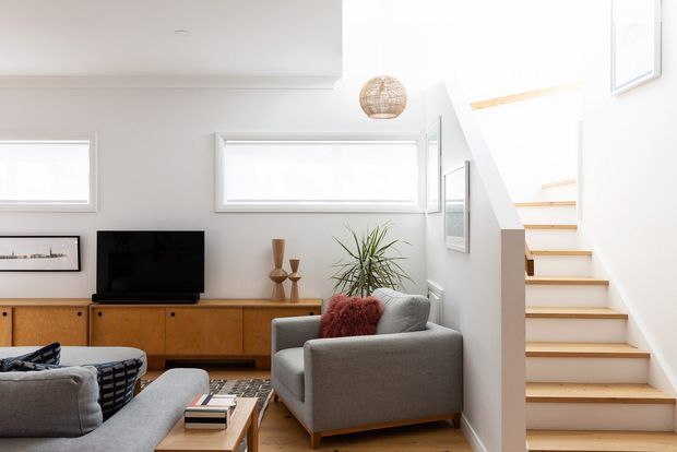 Bright, compact contemporary living room with a grey sofa and armchair, timber media cabinetry, framed artwork, indoor plants, and a timber coffee table. A white staircase with timber treads rises beside the seating area, and high horizontal windows provide abundant natural light.
