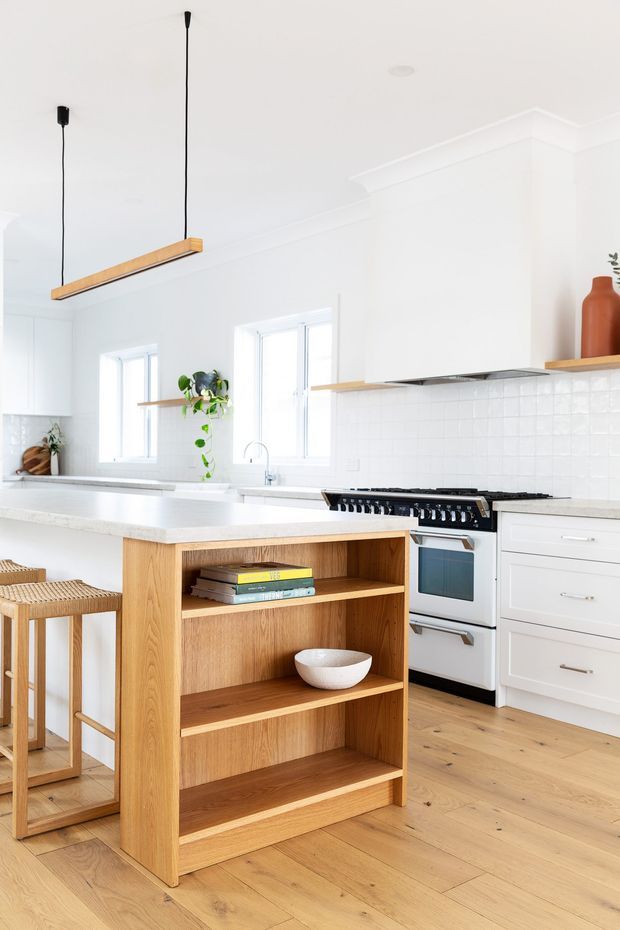 Bright, airy kitchen featuring a large white island with an integrated open timber shelving unit, white shaker-style cabinetry, pale timber flooring, white tiled splashback, and a freestanding stainless-steel range cooker. Linear timber pendant lights, natural wood bar stools, and a few restrained decorative elements add warmth to the predominantly white scheme.