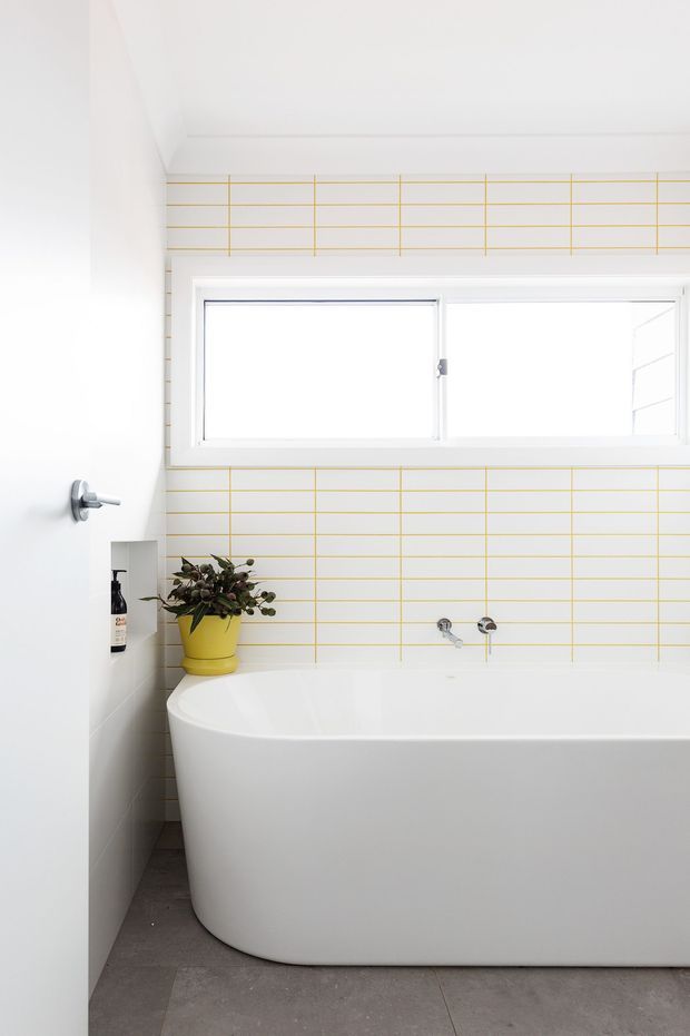 Bright, compact contemporary bathroom featuring a freestanding white bathtub, white wall tiles with a fine yellow grid pattern, a wide horizontal window, grey floor tiles, and a small yellow planter with greenery. The space has a clean, playful, high-contrast finish.
