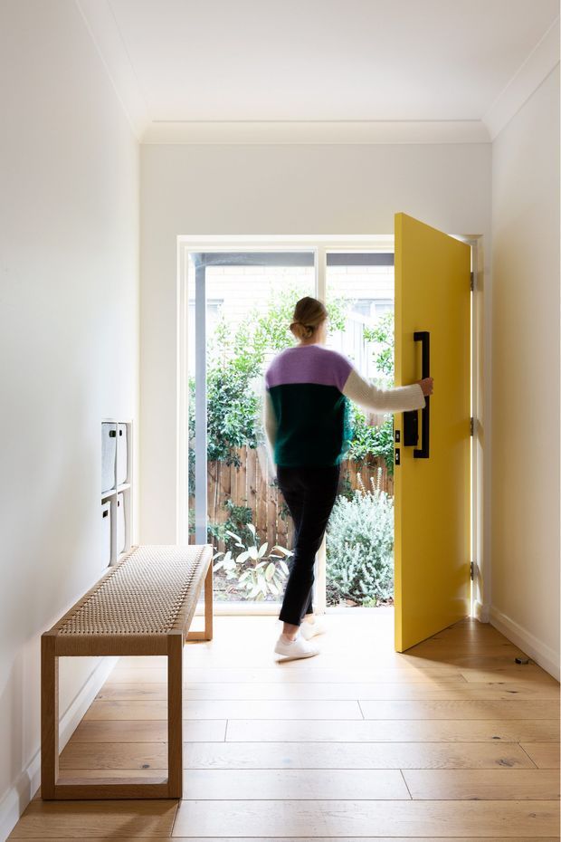 Bright residential entryway with pale timber flooring, white walls and ceiling, a woven timber bench, and a vivid mustard-yellow front door opening to a small planted courtyard. A person is exiting through the doorway, and a large glazed panel beside the door brings in natural light.