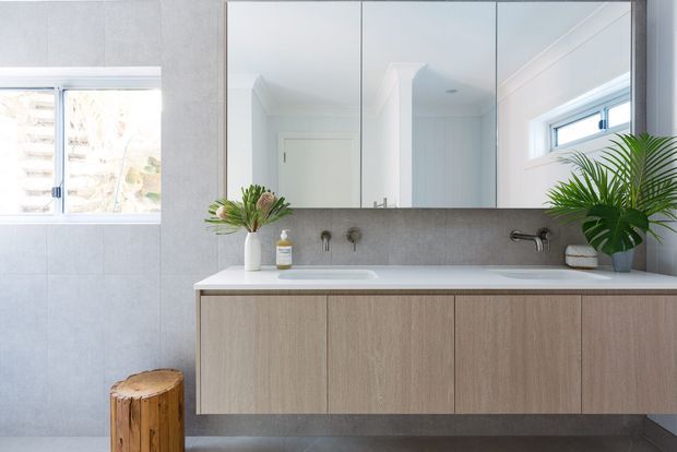 Bright contemporary bathroom featuring a wall-mounted timber-look vanity with a white integrated basin, large frameless mirrored cabinet, grey tiled walls and floor, wall-mounted brushed metal tapware, and high horizontal windows. A potted palm and small natural timber stool add organic accents.