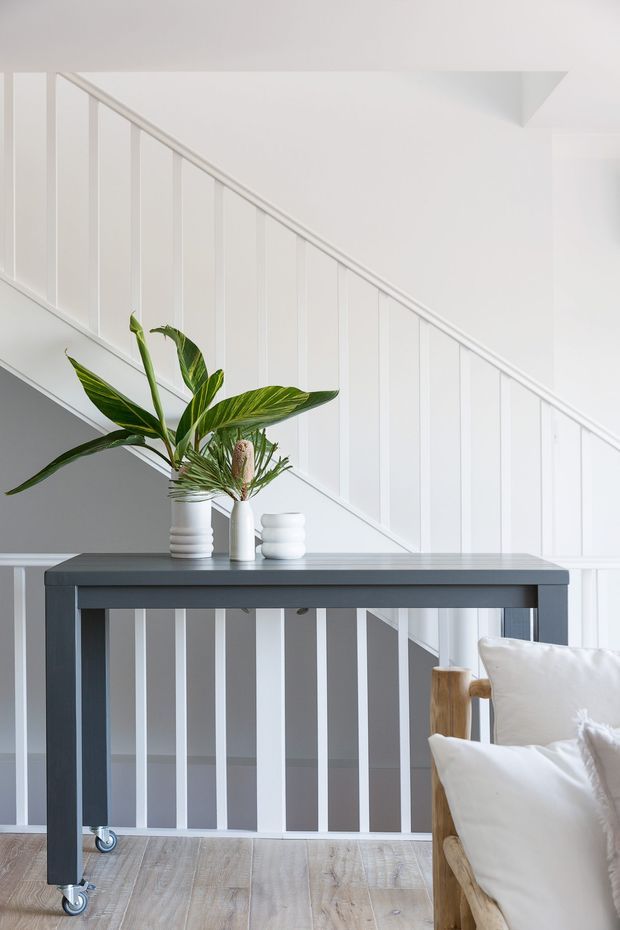 Bright, minimalist interior view of a white staircase with vertical balustrade panels and a slim handrail. A dark grey console table with caster wheels stands in front, topped with white ceramic vessels and green tropical foliage. Pale timber flooring and part of a light upholstered armchair are visible at the right edge.