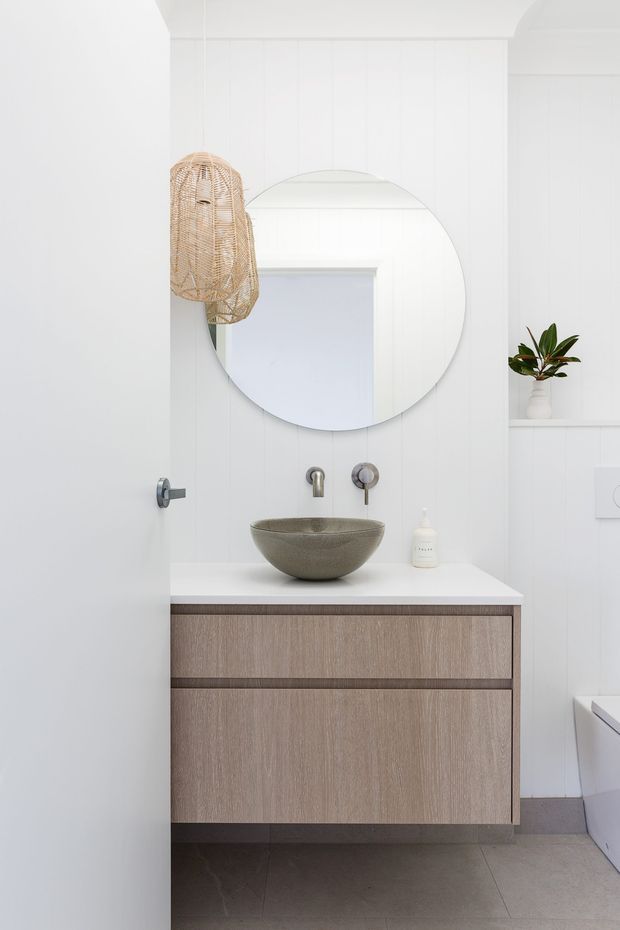 A bright, compact contemporary bathroom viewed through a partially open door. The room features white vertical wall panelling, a floating timber-toned vanity with integrated drawers, a white benchtop, a round vessel basin, wall-mounted brushed metal tapware, and a large round mirror. A woven pendant light and a small plant add natural texture and warmth. Grey floor tiles and a partial bathtub are visible.