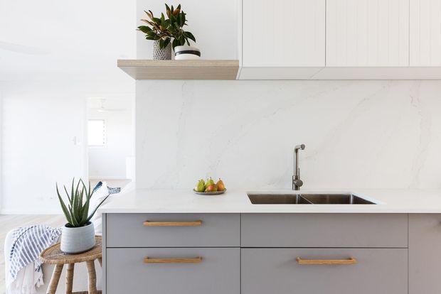Bright contemporary kitchen with pale grey flat-panel cabinetry, warm timber handles, a white marble-look splashback and benchtop, and a stainless steel undermount sink with mixer tap. Open shelving displays potted plants, while a small timber stool and plant sit at the side.