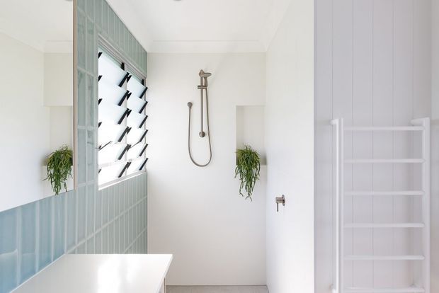 Bright, minimalist bathroom with a walk-in shower, white walls, pale blue-green vertical wall tiles, louvre window, wall-mounted shower hardware, hanging plants, and a white heated towel rail. A white vanity or basin edge is visible in the foreground.