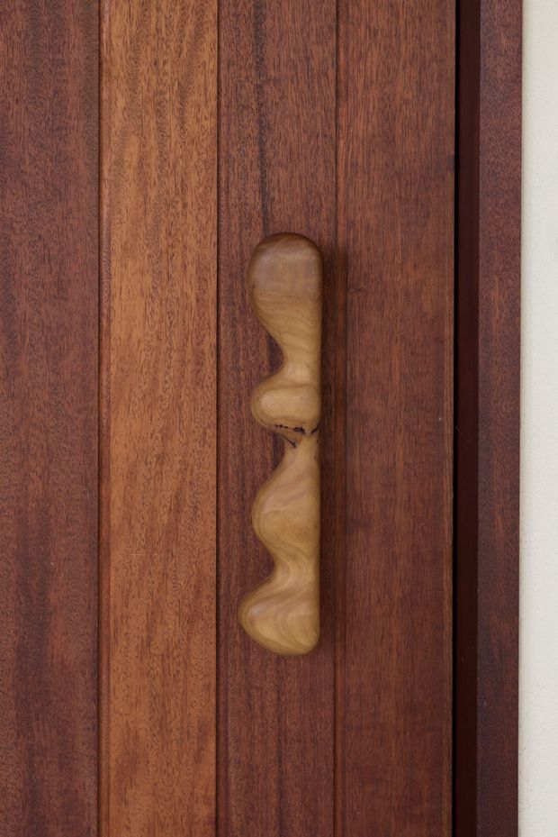 Close-up detail of a dark, richly grained timber door or wall panel with vertical boards and a sculptural organic timber handle. The composition focuses on the warm wood grain, joinery lines, and tactile bespoke hardware against a narrow light-coloured wall edge.