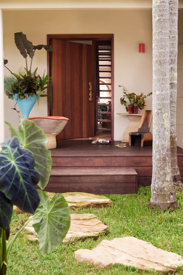 A tropical residential entry viewed from the garden, featuring a deep dark timber porch with broad steps, a richly stained timber front door, and a narrow glazed louvre or slatted sidelight. The entry is framed by warm beige rendered walls, a sculptural timber console with a potted plant, a woven lounge chair, and abundant tropical foliage. Irregular natural stone stepping slabs lead across the lawn toward the porch, with a palm trunk visible at the right.