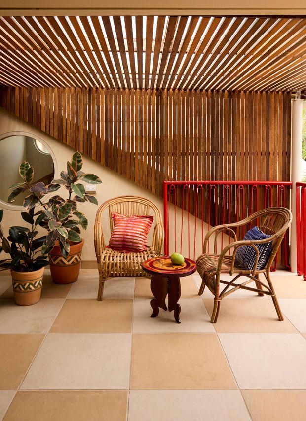 A warm, sheltered outdoor or semi-outdoor lounge with two woven rattan chairs, a small dark wood side table, potted plants, and a red-painted metal balustrade. The space features a dramatic timber slatted ceiling and matching vertical timber privacy screen, with large beige square floor tiles and a rounded wall mirror. The colourful cushions and plants create a relaxed, tropical-inspired atmosphere.