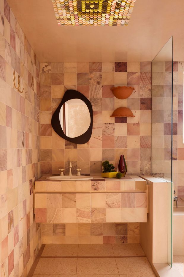 A compact, richly detailed bathroom vanity area clad in a patchwork of warm pink, terracotta, beige, and cream stone or ceramic tiles. A floating timber-toned vanity with integrated tiled drawer fronts supports a white basin and vintage-style tapware. The space features an organic black-framed oval mirror, sculptural wall decor, colourful accessories, a decorative gridded ceiling light, and a frameless glass shower screen at right. The overall effect is warm, playful, artistic, and highly layered.
