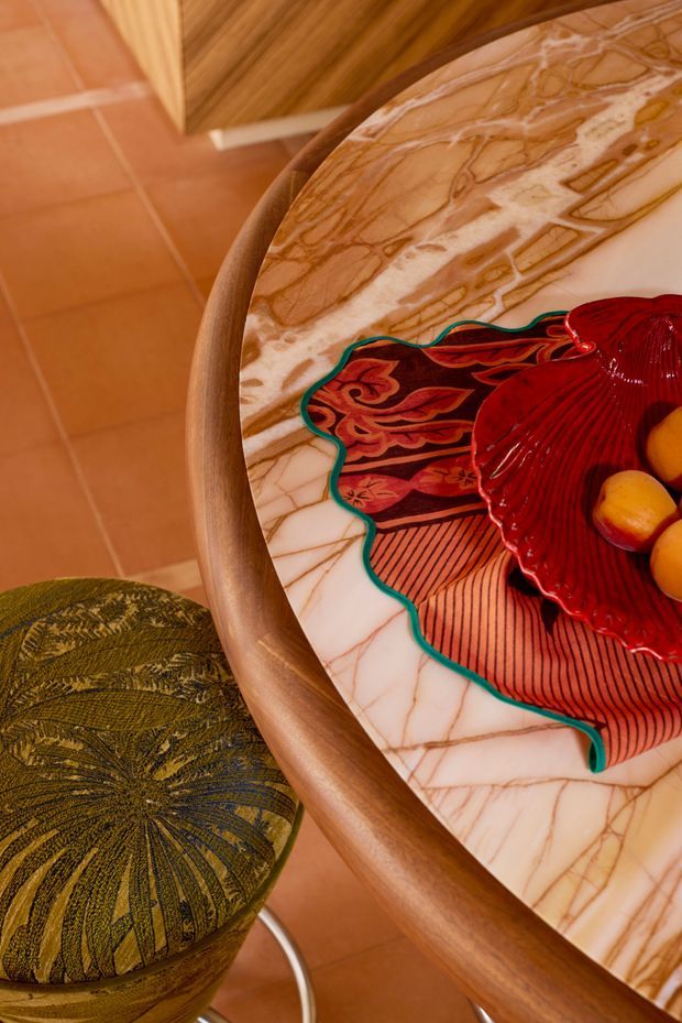 Close-up view of a richly styled dining area, focused on a round table with a veined marble tabletop and rounded timber edge. A vivid red glass fruit bowl and patterned decorative textile sit on the table, with a green patterned upholstered stool and terracotta tiled flooring visible nearby. The composition has a warm, eclectic and retro-inspired character.