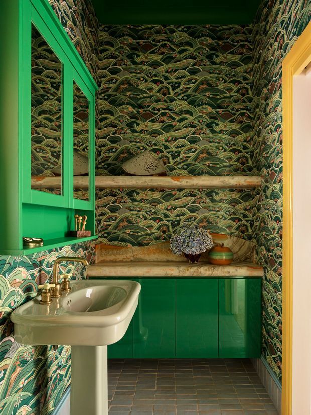 A compact, highly decorative bathroom featuring dark green, wave-patterned wallpaper on all walls, glossy emerald-green built-in cabinetry and open shelving, a vintage-style cream pedestal basin with brass taps, and a weathered stone or marble countertop and shelf. The dark tiled floor and yellow-painted door trim contrast with the saturated green palette. A small arrangement of decorative objects sits on the vanity, creating an eclectic, richly layered interior.