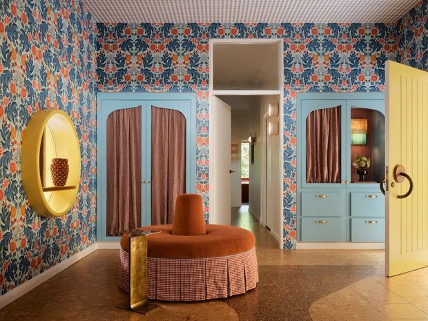 A highly decorative residential entryway or hallway with bold floral wallpaper covering the walls and ceiling, turquoise built-in cabinetry and wardrobes, and a bright yellow open door. A sculptural rust-orange upholstered seat with a checked skirt sits centrally, accompanied by a gold-toned side table. The space has a playful, richly patterned maximalist character, with a corridor leading to a brighter room beyond.