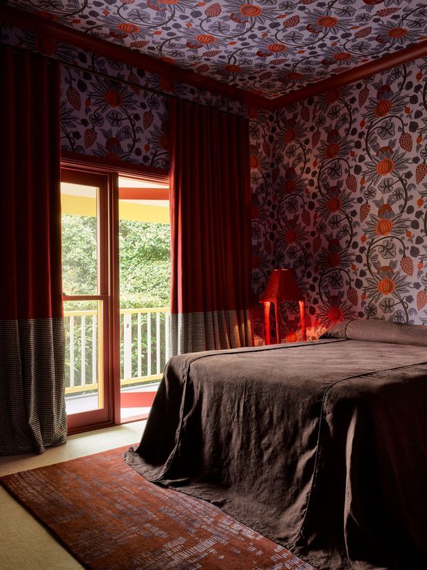A richly layered, moody bedroom featuring dark floral botanical wallpaper across the walls and ceiling, deep red curtains with checked lower panels, and a dark draped bed. A red bedside lamp sits beside the bed, while a glazed balcony door brings in daylight and overlooks greenery. The room has an eclectic, dramatic and highly patterned interior with warm timber framing and a patterned rug.