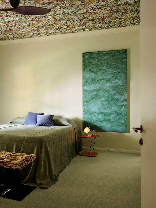 A moody, richly layered bedroom with a low-profile bed dressed in olive-green bedding and lavender cushions. Pale warm walls contrast with a large textured green feature artwork or wall panel, while a highly patterned floral ceiling adds a bold decorative element. A dark sculptural ceiling fan, slim side table, small lamp, and patterned upholstered bench complete the room.