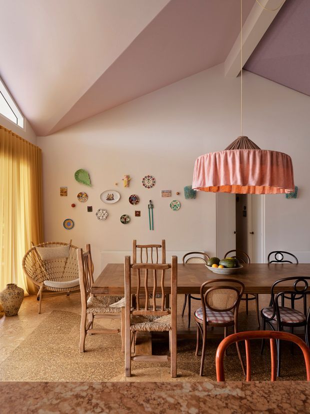 A warm, eclectic dining room with a large timber dining table surrounded by mismatched wooden and bentwood chairs. A woven lounge chair sits beside full-height mustard curtains, while a decorative pink pleated pendant hangs over the table. The pale walls display a collection of small decorative plates and artworks, and a faceted stone or terrazzo floor adds texture. The room has a distinctive angled, high ceiling with a small high-level window and soft pink-toned ceiling planes.