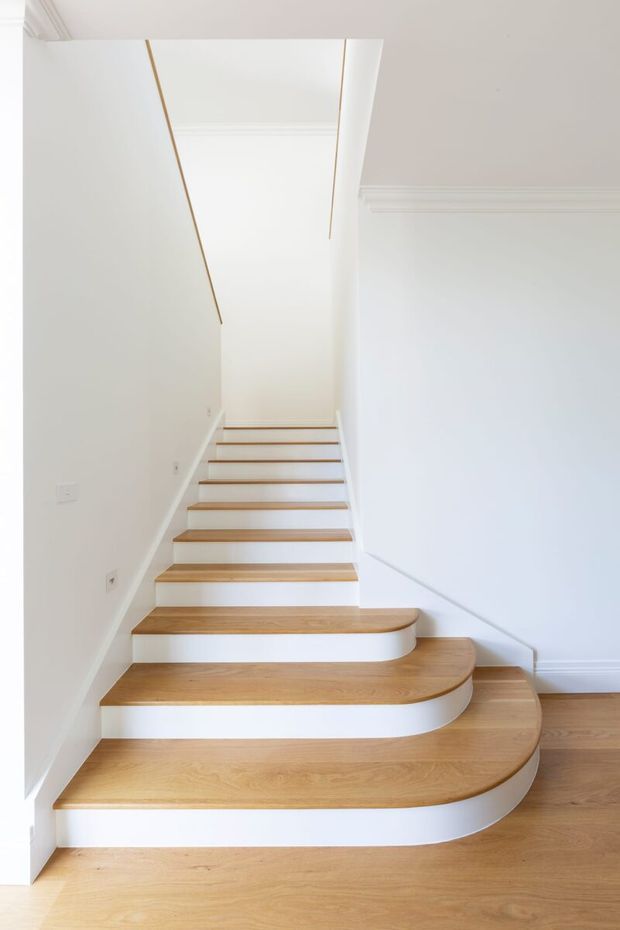 Bright minimalist interior view of a staircase with broad, curved timber treads and white painted risers. The stair turns through a series of rounded steps, with white walls, simple moulded cornices, and a light timber floor creating a clean, refined appearance.