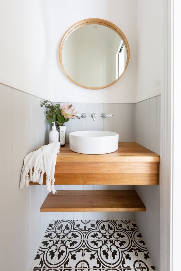Compact, bright bathroom vanity nook with a round timber-framed mirror, white vessel basin, wall-mounted chrome tapware, timber floating vanity and shelf, pale vertical wall panelling, and patterned black-and-white floor tiles. A small floral arrangement and folded textile add softness.