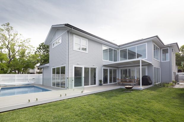 Exterior rear view of a two-storey contemporary home with light grey horizontal weatherboard cladding, extensive white-framed glazing, covered outdoor terrace, and a swimming pool set beside a landscaped lawn.