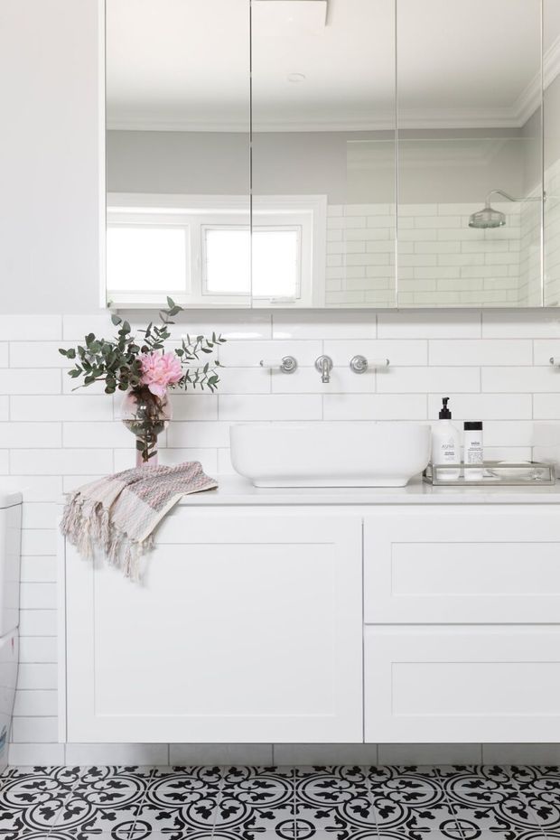Bright, white bathroom vanity with a freestanding basin, wall-mounted chrome tapware, mirrored upper cabinets, white subway-tile walls, and a patterned black-and-white tiled floor. A shower is visible in the mirror reflection, with a small floral arrangement and folded throw adding a soft decorative touch.