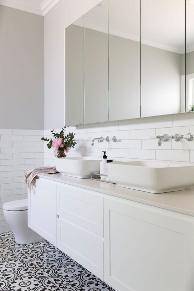 Bright, refined bathroom with a double vanity, two white vessel basins, wall-mounted chrome taps, mirrored upper cabinetry, white shaker-style joinery, white subway-tile walls, and an ornate black-and-white patterned floor tile. A toilet and floral arrangement are also visible.