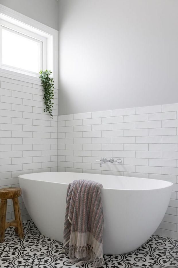 A bright, compact bathroom featuring a freestanding white bathtub, white subway-tile walls, patterned encaustic-style floor tiles, a high white-trimmed window, chrome wall-mounted bath fittings, and a small wooden stool. A fringed woven throw is draped over the tub, with a trailing green plant adding a natural accent.