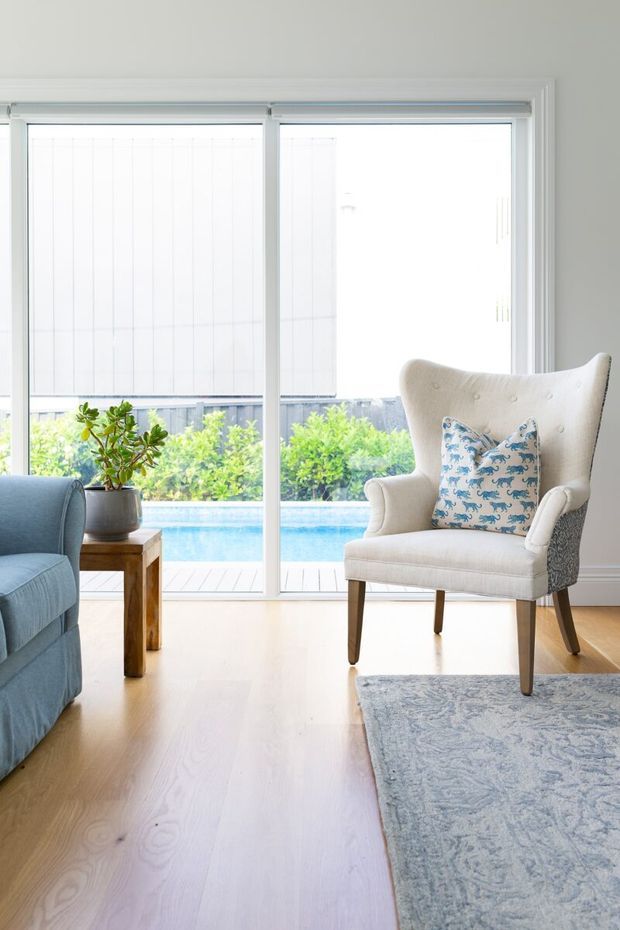 Bright, airy living room with large white-framed glazed sliding doors overlooking a backyard swimming pool. The room features pale timber flooring, a light blue sofa, a cream wingback armchair with a blue patterned cushion, a small timber side table, a potted plant, and a pale patterned rug.