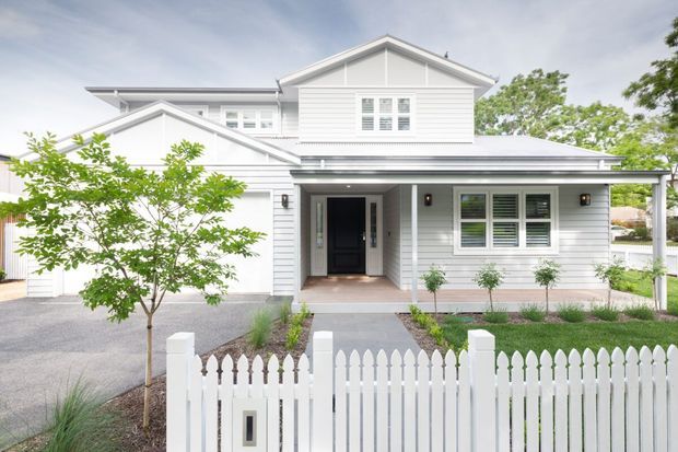 Front exterior view of a light-coloured, multi-level suburban residence with horizontal weatherboard cladding, a pitched gable roof, covered entry porch, white-framed windows with shutters, attached garage, landscaped garden, and white picket fencing.