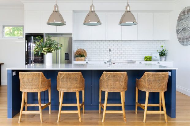 Bright, refined kitchen featuring a large navy island with a white benchtop and four woven timber bar stools. White shaker-style cabinetry, stainless-steel appliances, a white subway-tile splashback, pendant lights, and light timber flooring create a fresh, coastal-leaning look.