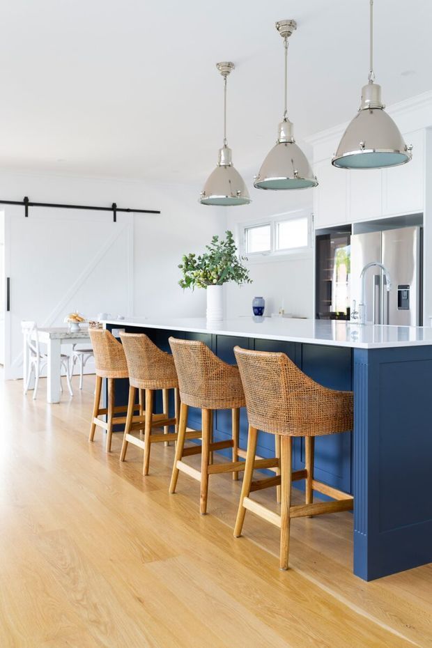 Bright, spacious open-plan kitchen and dining area with a large navy-blue island, white stone-look benchtop, four woven rattan bar stools, pale timber flooring, white cabinetry, and three oversized pendant lights. A white barn-style sliding door and dining table are visible in the background, creating a light coastal-inspired interior.