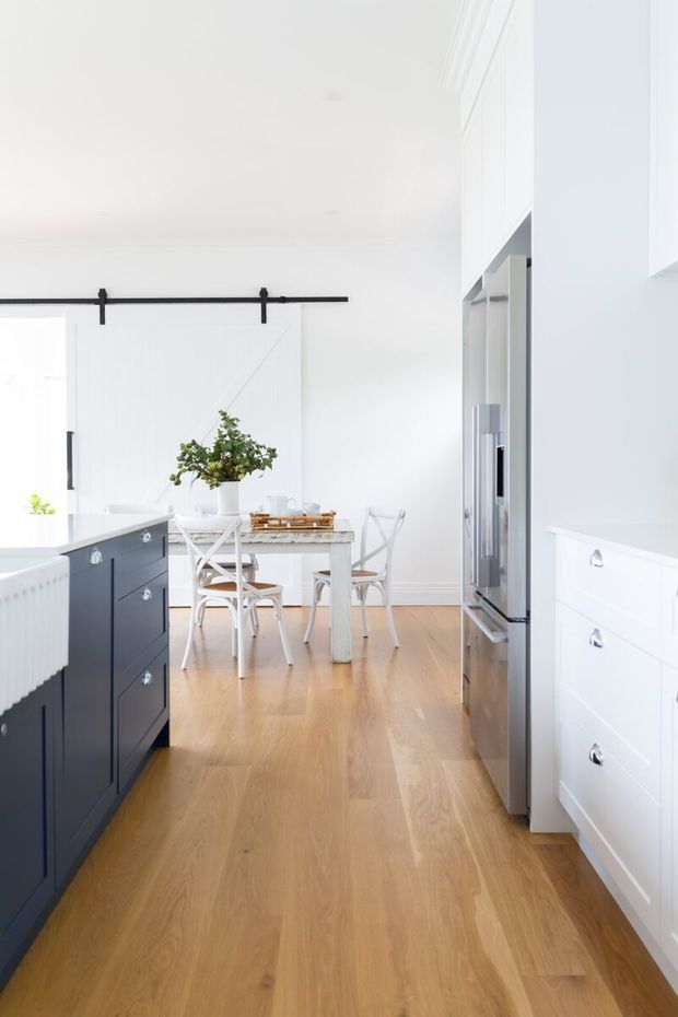 Bright, narrow kitchen with white walls and cabinetry, a dark navy island, stainless-steel appliances, and warm timber flooring. A small white dining table with cross-back chairs sits beyond the kitchen, separated by a white barn-style sliding door with black hardware. The room has a clean, airy contemporary farmhouse character.